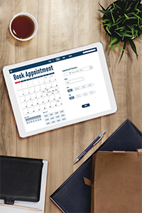 Tablet screen showing an online appointment booking system with calendar view, surrounded by a coffee cup and notepad on a wooden desk.